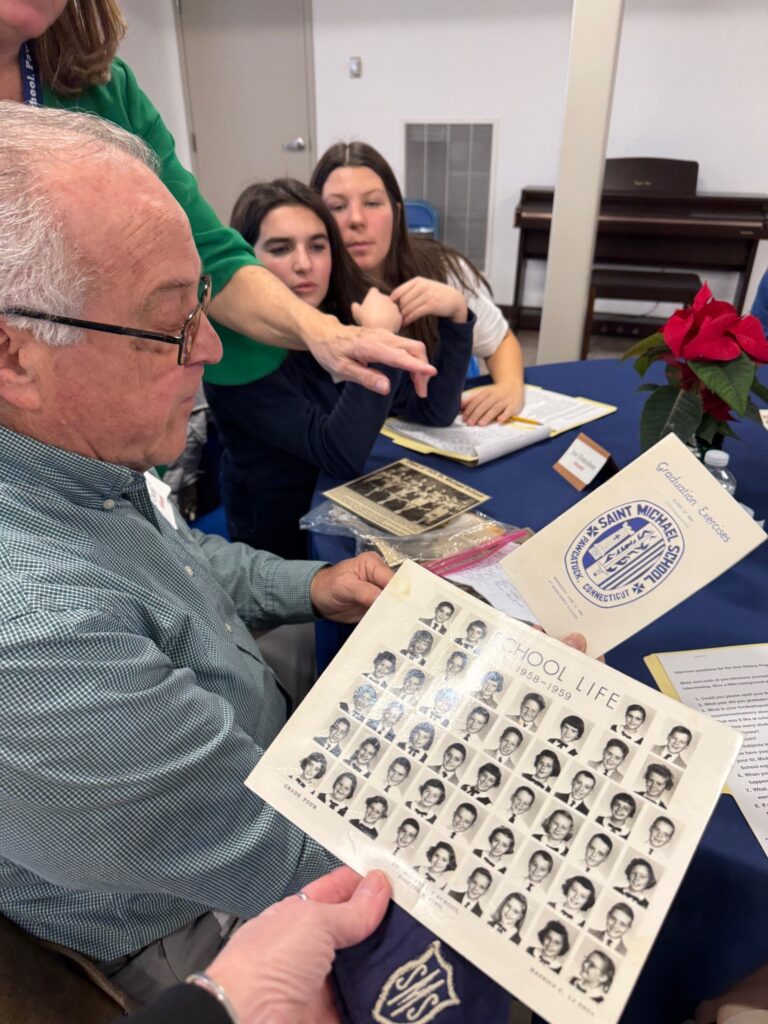 Man with gray hair and glasses looking at class photos with female students in the background, looking on