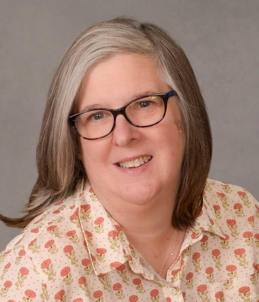 Smiling woman with light hair wearing dark framed glasses and a white shirt with pink flowers