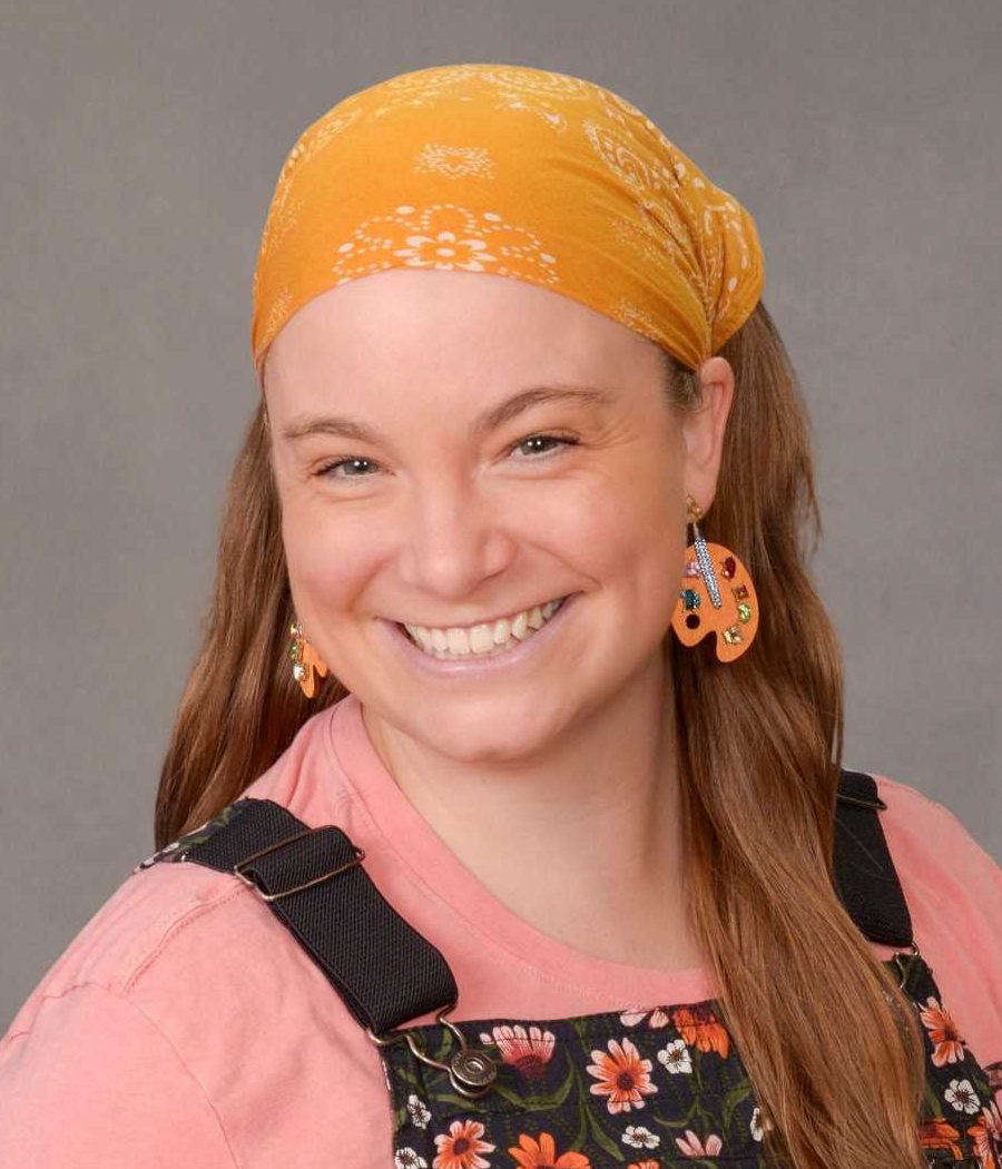 Smiling young woman with bright yellow hair bandana wearing artist palette earrings, dark blue flowered overalls and a pink shirt