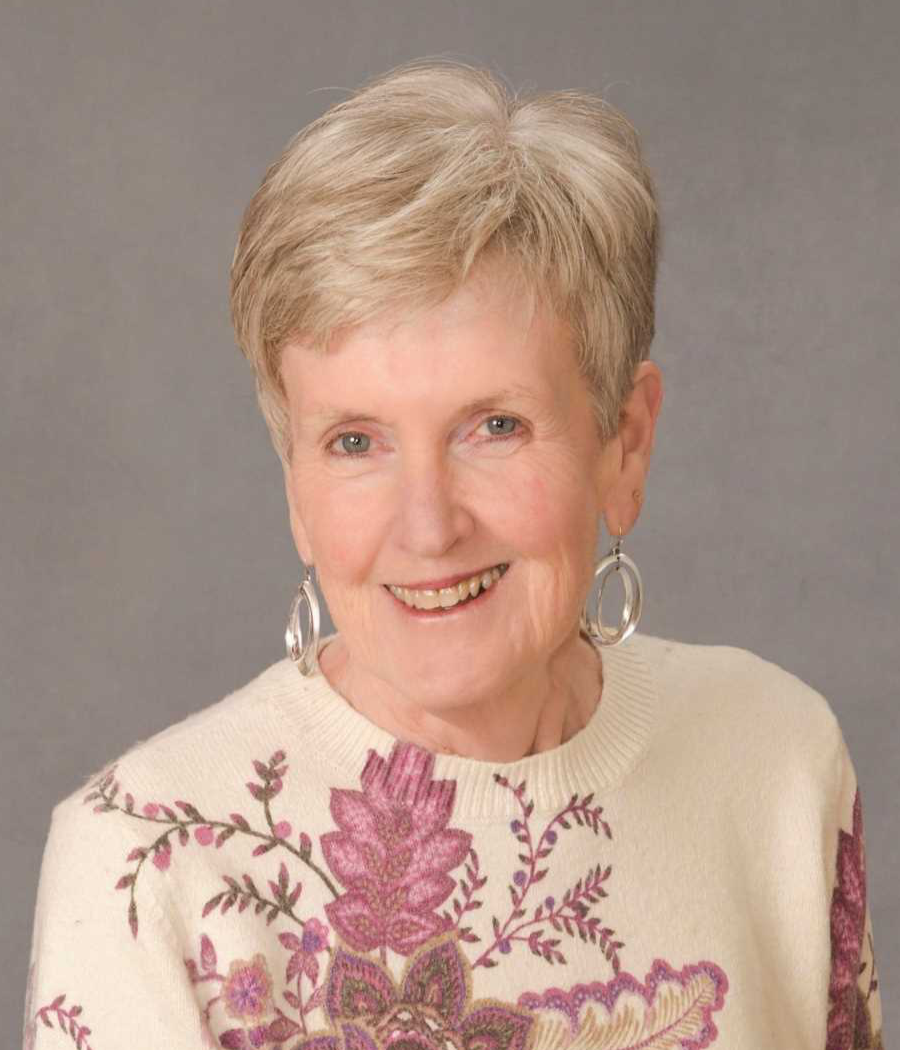 Smiling woman with short blonde hair wearing large silver earrings and a cream colored sweater with large pink flowers