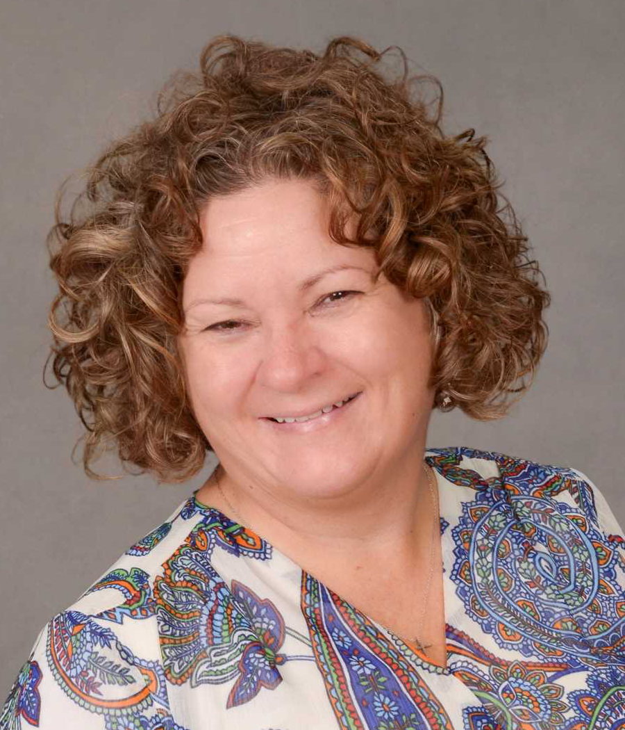 Smiling woman with short curly brown hair wearing a white shirt patterned with blue, green and orange