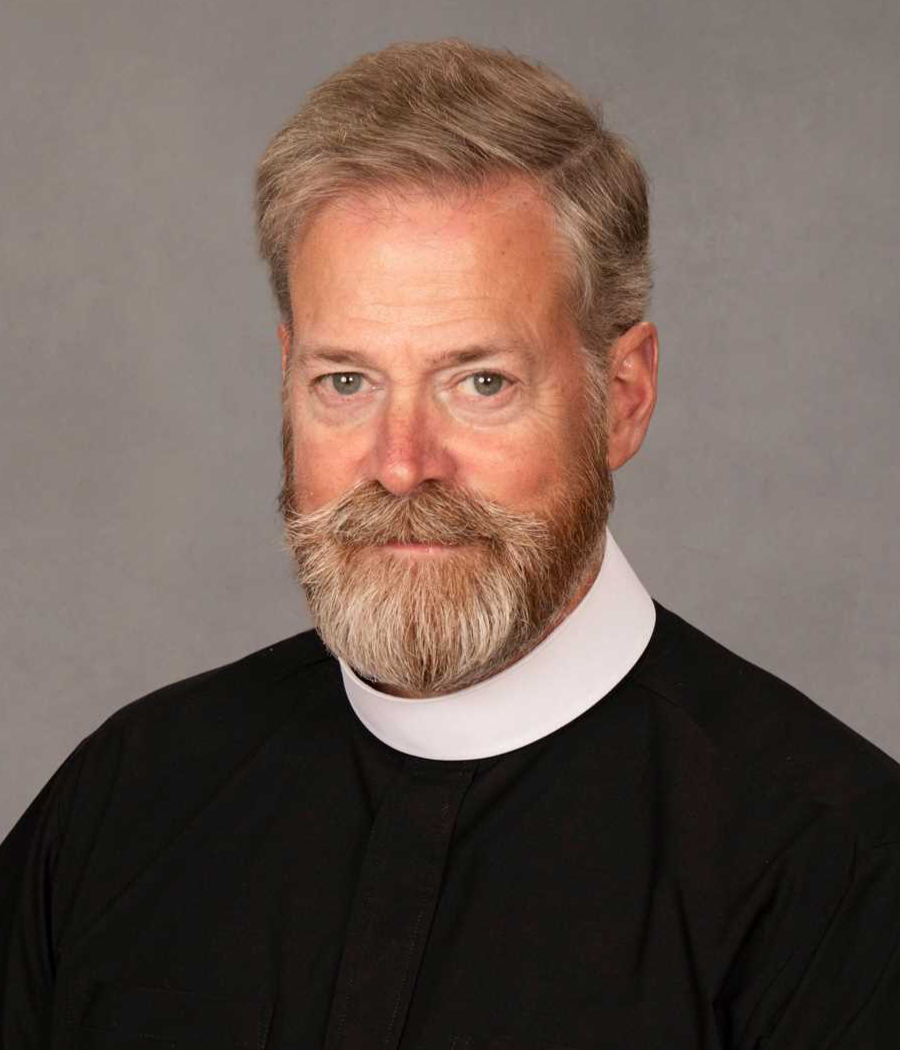 Smiling man with light brown hair and beard in a black shirt and clerical collar