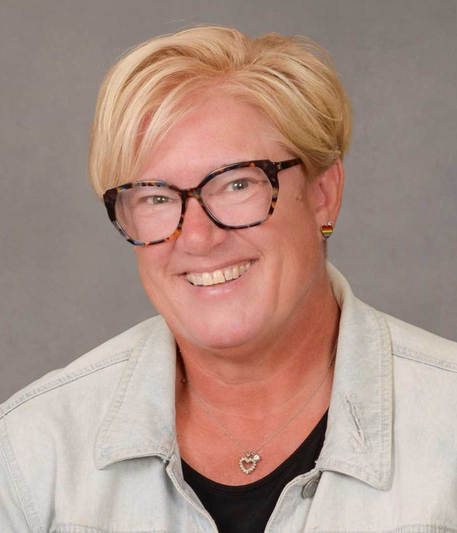Smiling woman with short blonde hair wearing dark glasses, a black shirt, and a light denim jacket