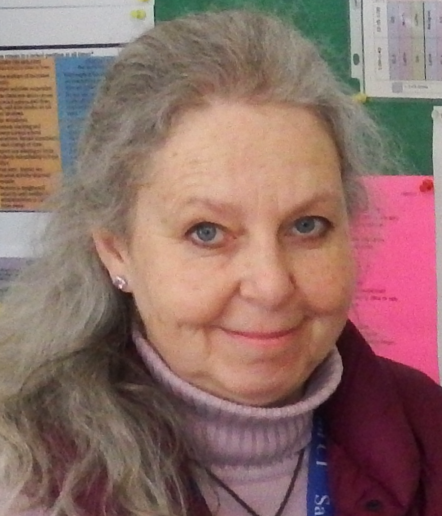 Woman with long, pulled-back light hair with a pink shirt and burgundy vest in front of a colorful bulletin board