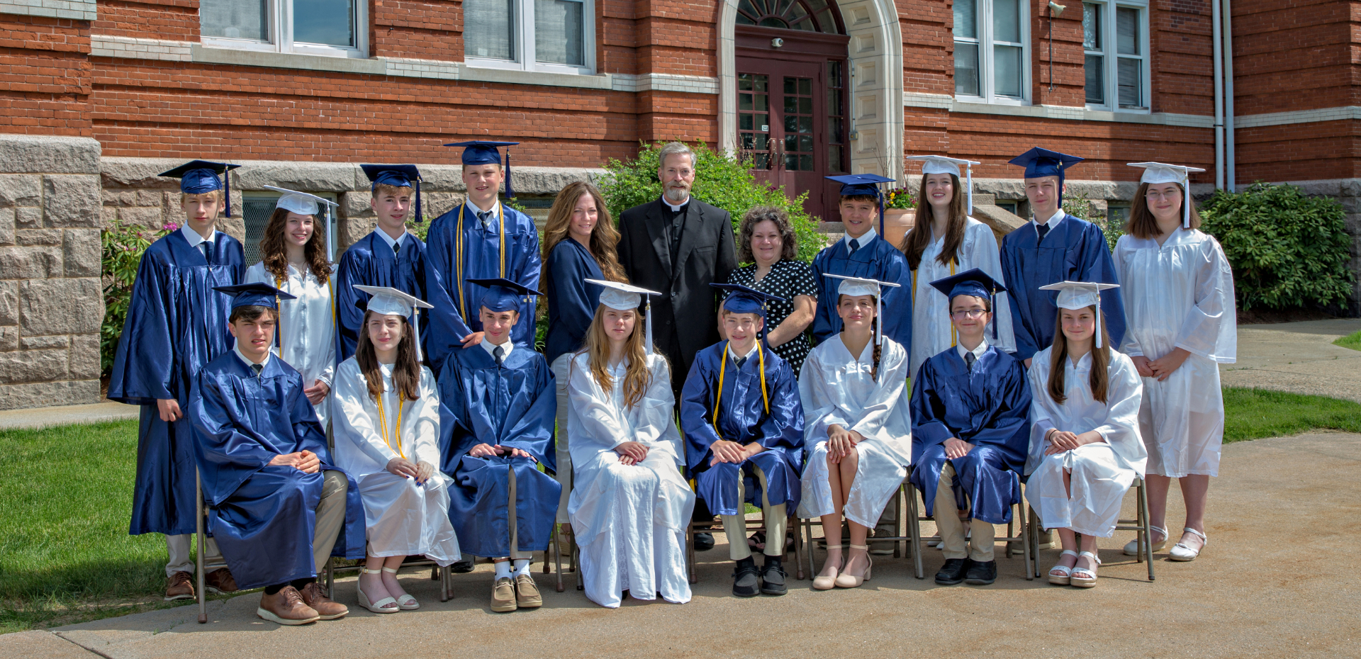 Photo from 2026 in front of a brick school building of a group young men in blue graduation caps and gowns and young women in white graduation caps and gowns with a man in a black priest suit and white collar and two women in dress clothes