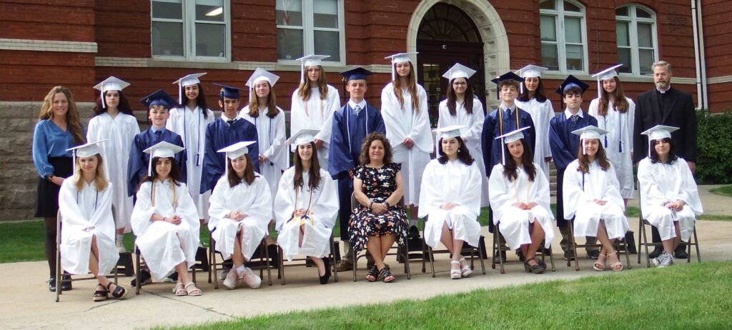 Graduates in blue and white caps and gowns with teachers and pastor in front of brick school building
