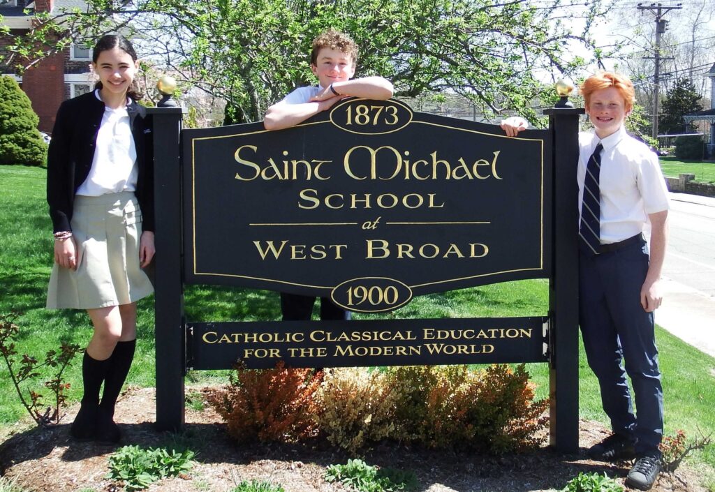 Young woman and two young men in school uniforms standing outside next to a black sign of Saint Michael School