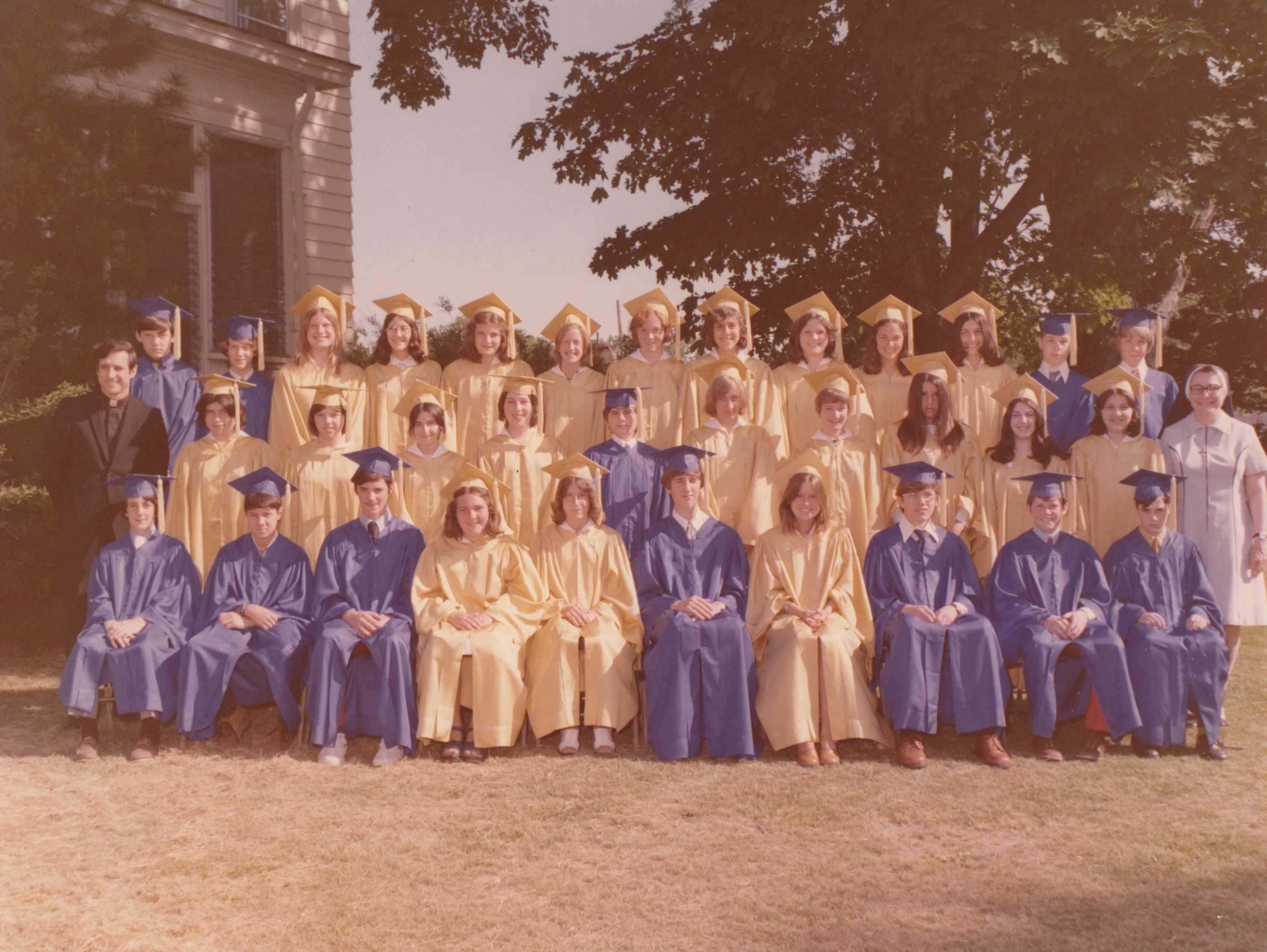 Photo from 1975 take in front of a building and large tree of a group young men in blue graduation caps and gowns and young women in yellow graduation caps and gowns with a man in a black priest suit and white collar and a woman in a white nun's habit and dark veil