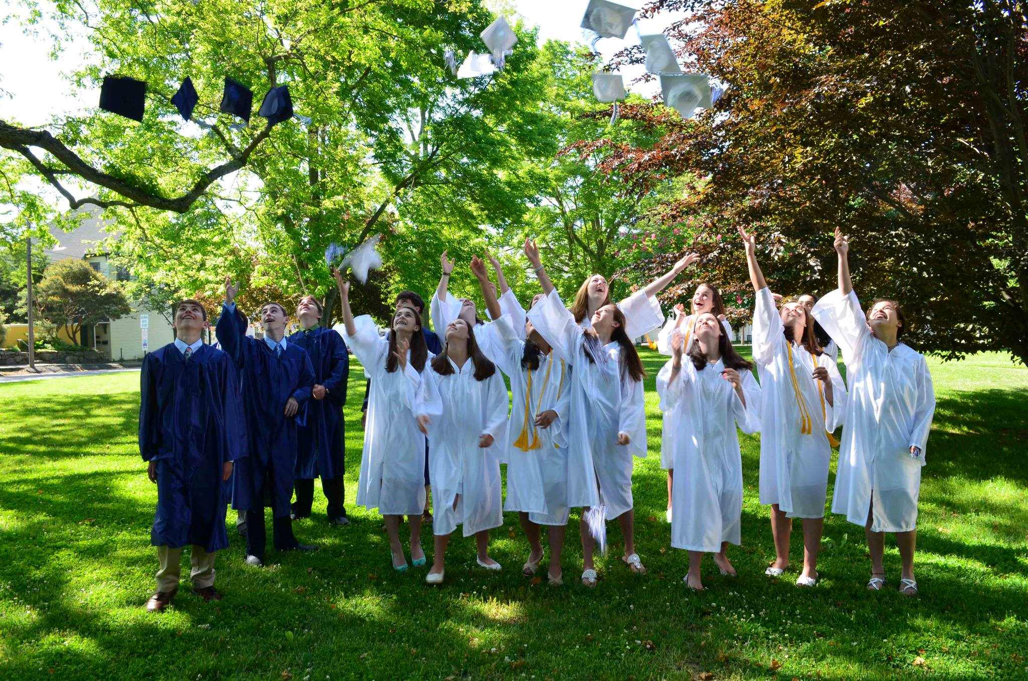 Photo from 2013 taken in front of trees of a group young men in blue graduation gowns and young women in white graduation gowns, throwing their caps in the air