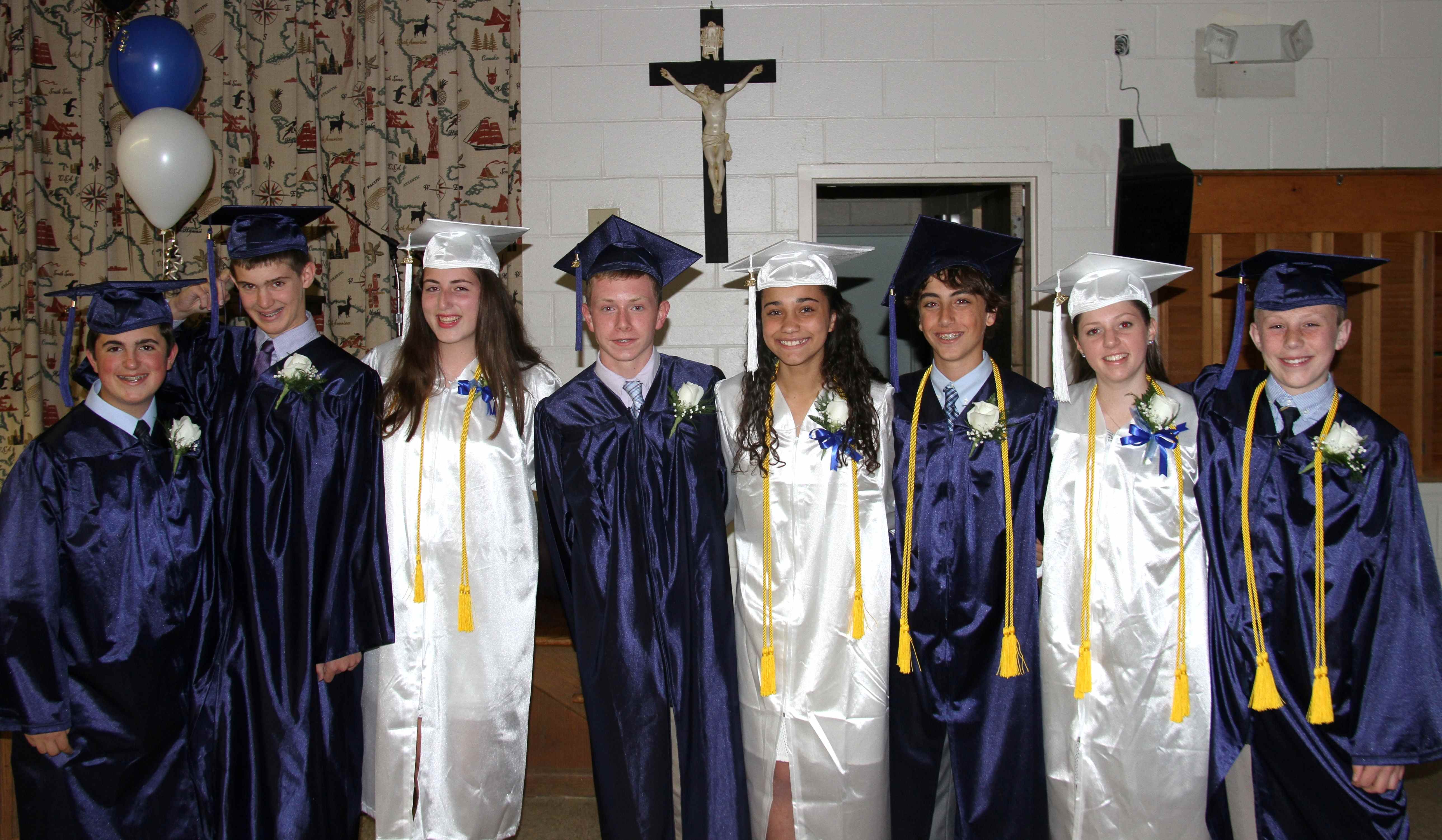 Photo from 2015 in front of a large crucifix of a group young men in blue graduation caps and gowns and young women in white graduation caps and gowns