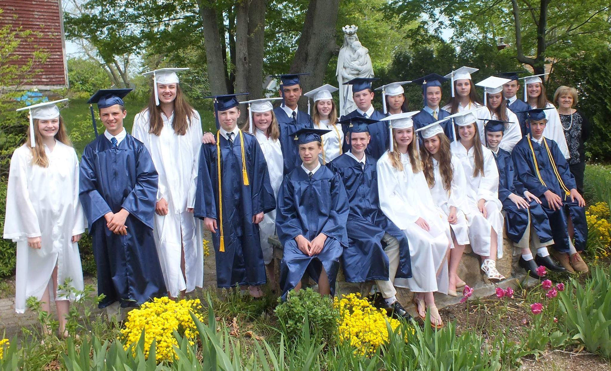Photo from 2016 in front of a statue of the Blessed Mother of a group young men in blue graduation caps and gowns and young women in white graduation caps and gowns with a woman in dress clothes