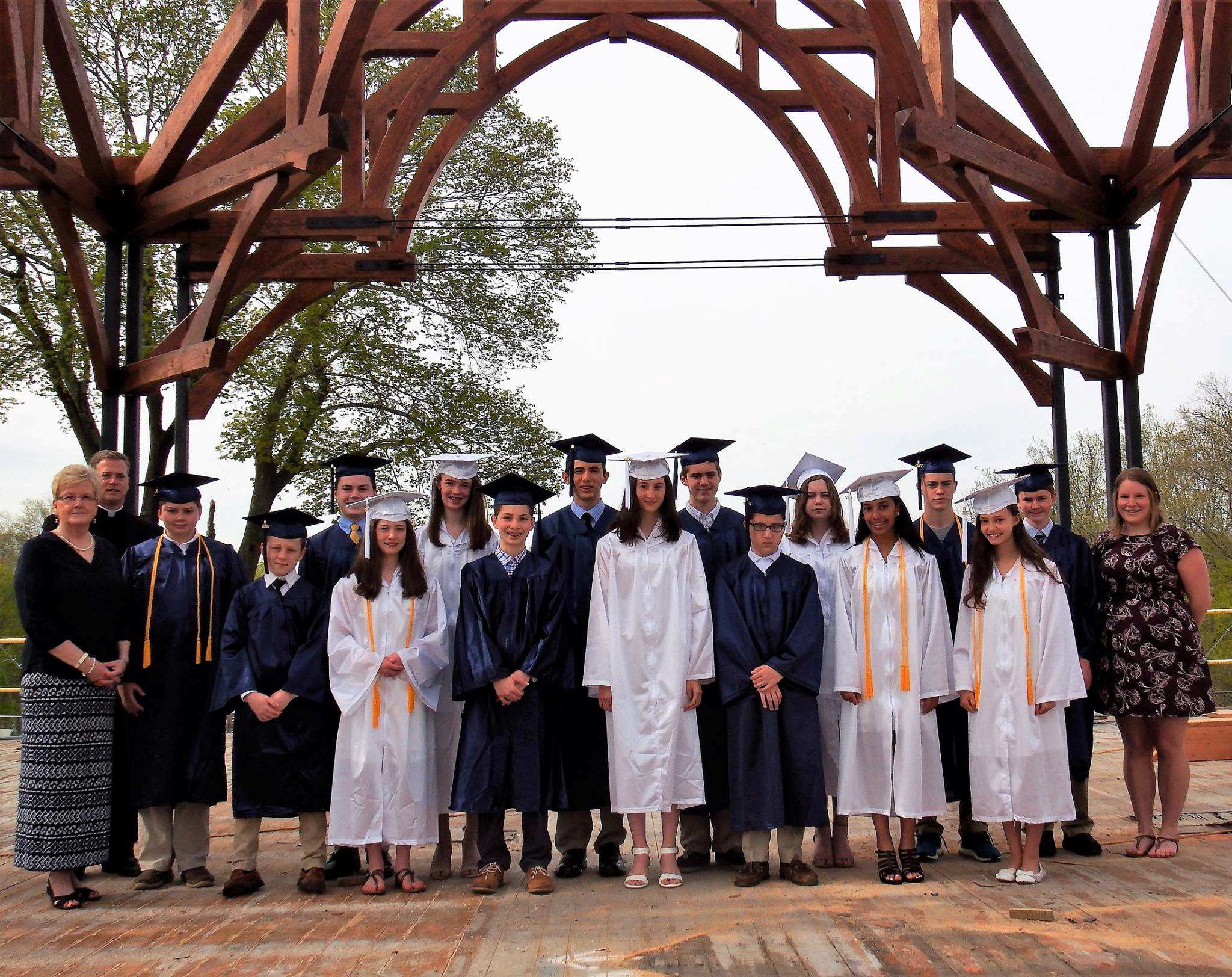 Photo from 2017 in front of a large overhead wooden structure of a group young men in blue graduation caps and gowns and young women in white graduation caps and gowns with a man in a black priest suit and white collar and two women in dresses
