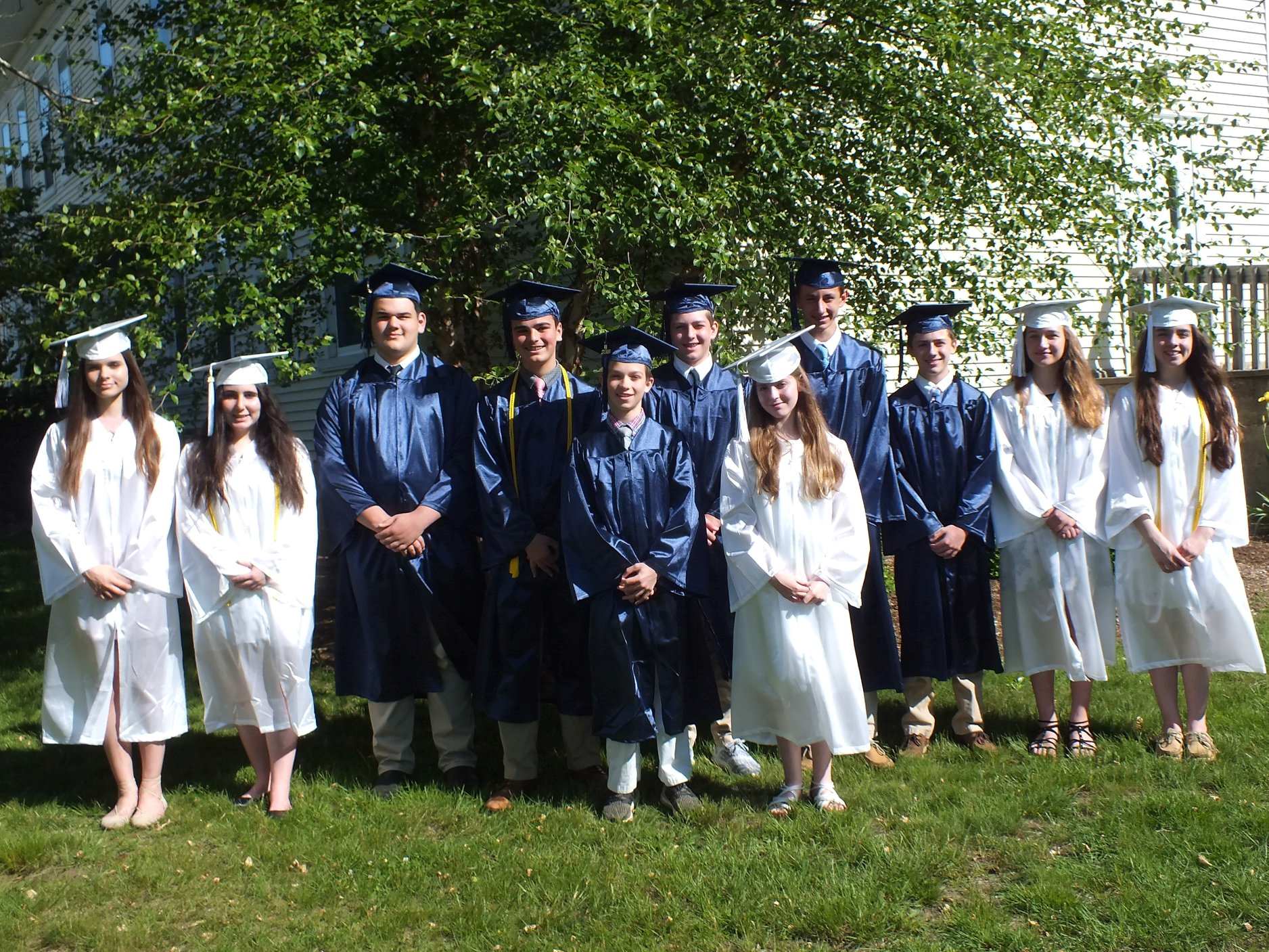 Photo from 2018 in front of a large tree of a group young men in blue graduation caps and gowns and young women in white graduation caps and gowns