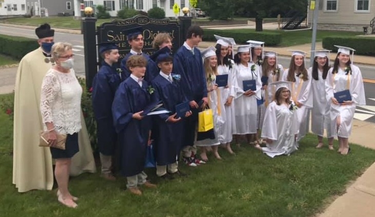 Photo from 2020 in front of a large black church sign of a group young men in blue graduation caps and gowns and young women in white graduation caps and gowns with a man in a white priest cassock and a woman in a black and white dress