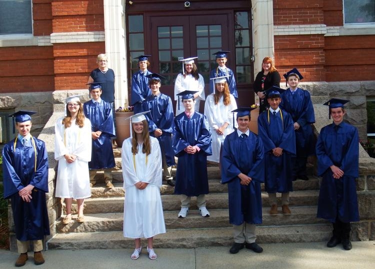 Photo from 2021 in front of a brick school building of a group young men in blue graduation caps and gowns and young women in white graduation caps and gowns with two women in dress clothes