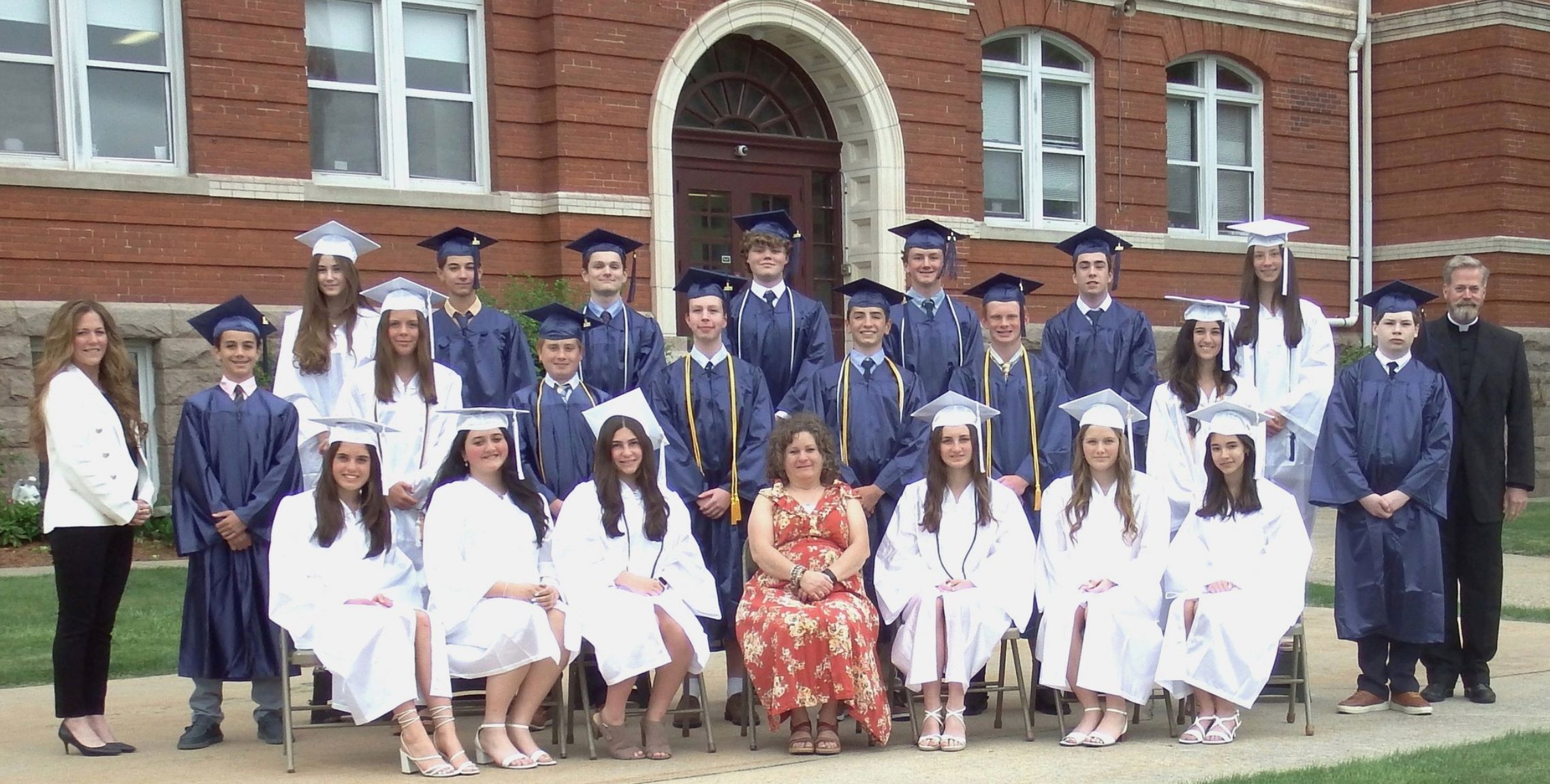 Photo from 2025 in front of a brick school building of a group young men in blue graduation caps and gowns and young women in white graduation caps and gowns with a man in a black priest suit and white collar and two women in dress clothes