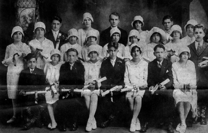 Black and white photo from 1928 of young women in white dresses with white hats and young men in dark suits