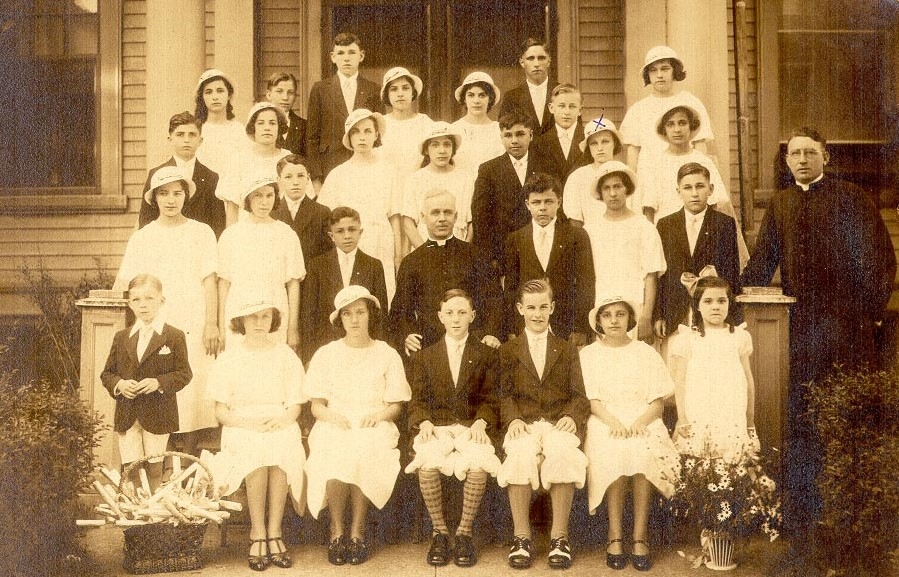 Sepia tone photo in front of a building from 1933 of a group of young women in white dresses and white hats with young men in dark suits with a man in a priest's cassock with a white collar