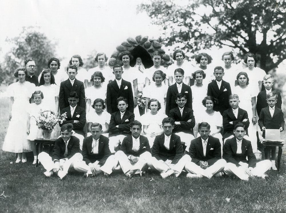 Black and white photo in front of outdoor grotto from 1934 of a group of young women in white dresses and young men in dark suits with a man in a priest's cassock and white collar
