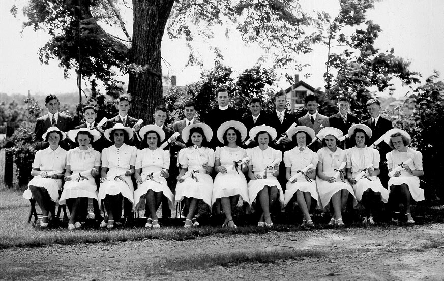 Black and white photo in front of a tree from 1940 of young women in white dresses and white hats and young men with dark suits and a man in priest's cassock and white collar