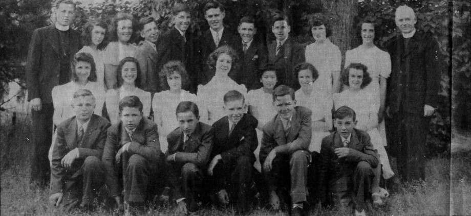 Black and white photo from 1942 of young women in white dresses and young men in dark suits with two men in priest's cassocks and white collars