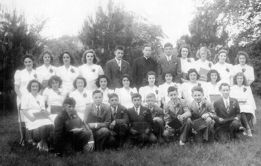 Black and white photo from 1943 taken outdoors of young women in white dresses and young men in dark suits with a man in a priest's cassock and white collar