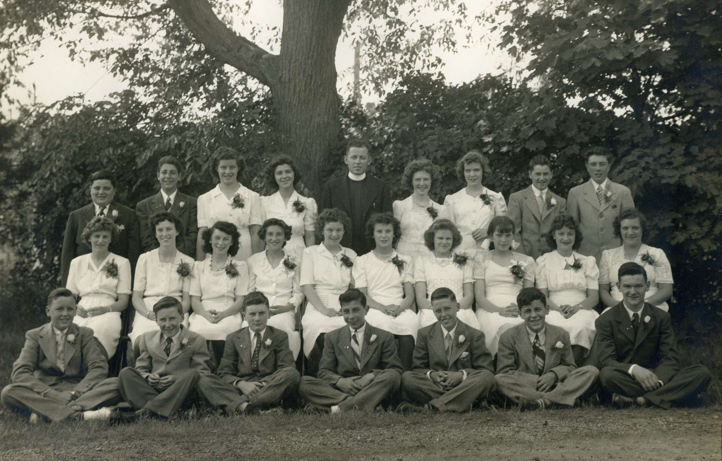 Black and white photo in front of a large tree from 1944 of a group of young women in white dresses and young men in dark suits with a man in a priest's cassock with a white collar