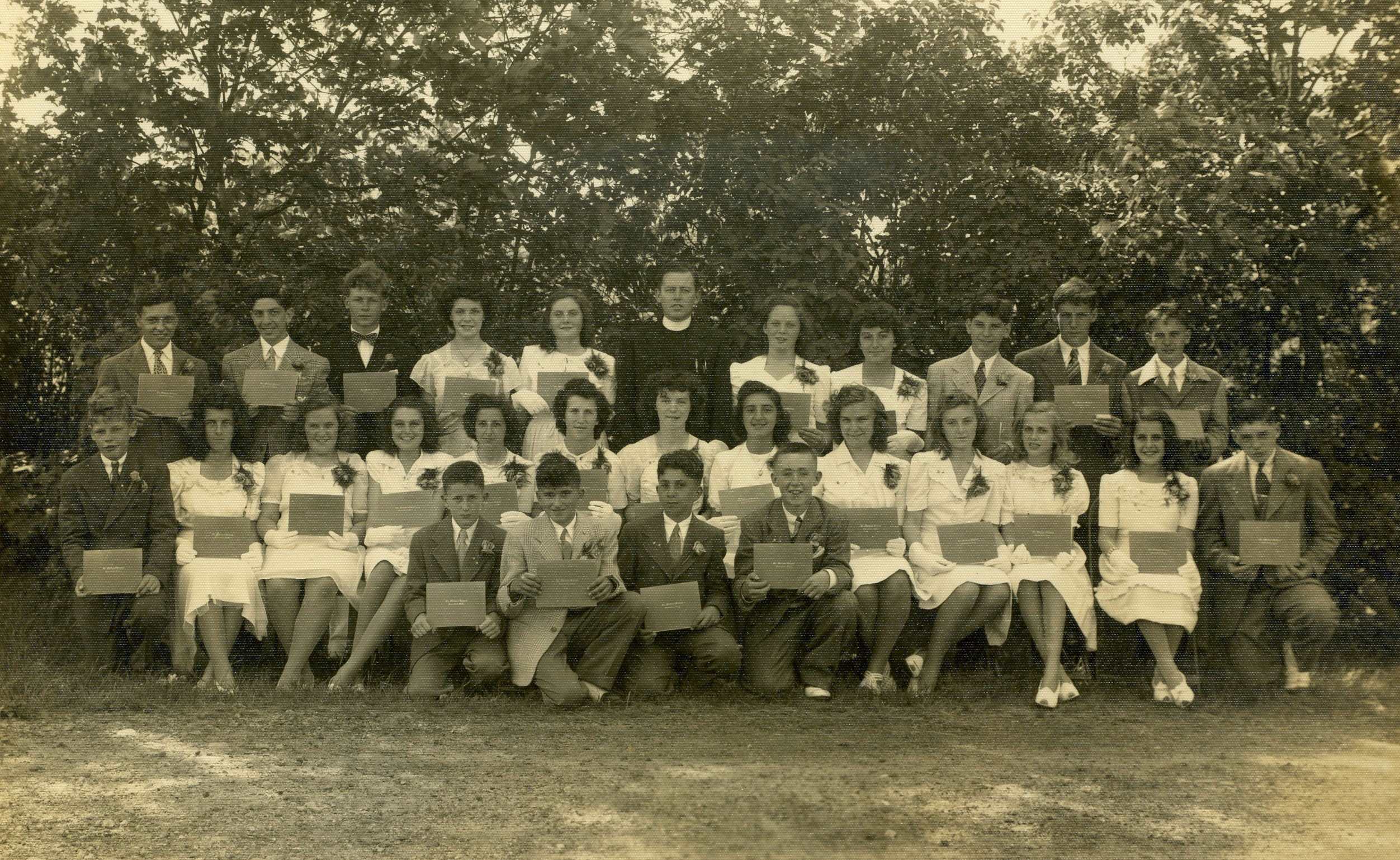 Black and white photo in front of a large tree from 1945 of a group of young women in white dresses and young men in dark suits with a man in a priest's cassock and white collar