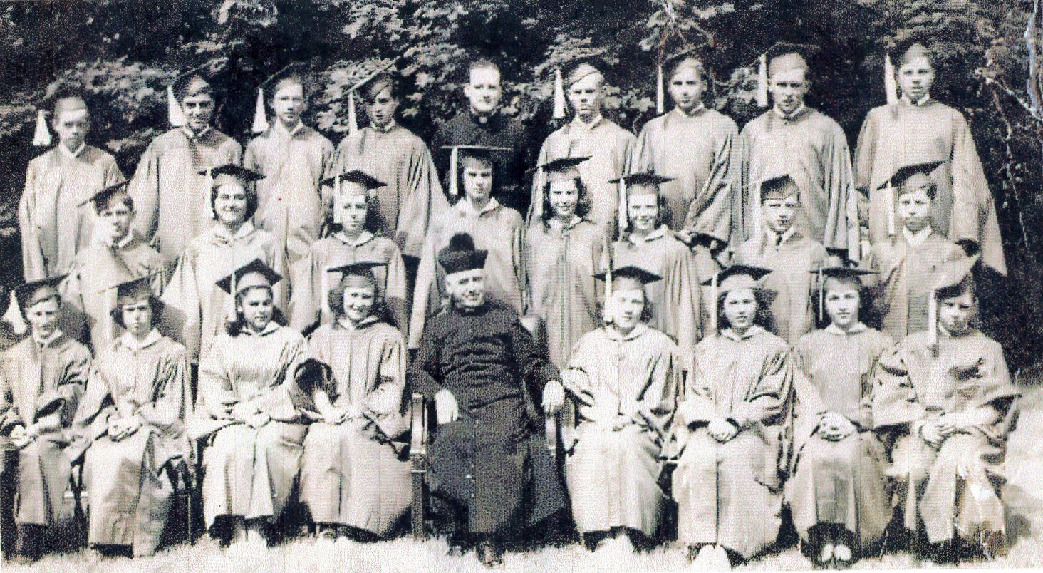 Black and white photo taken outdoors from 1948 of a group of young men and women in graduation robes and caps with a man in a priest's cassock and white collar and black hat