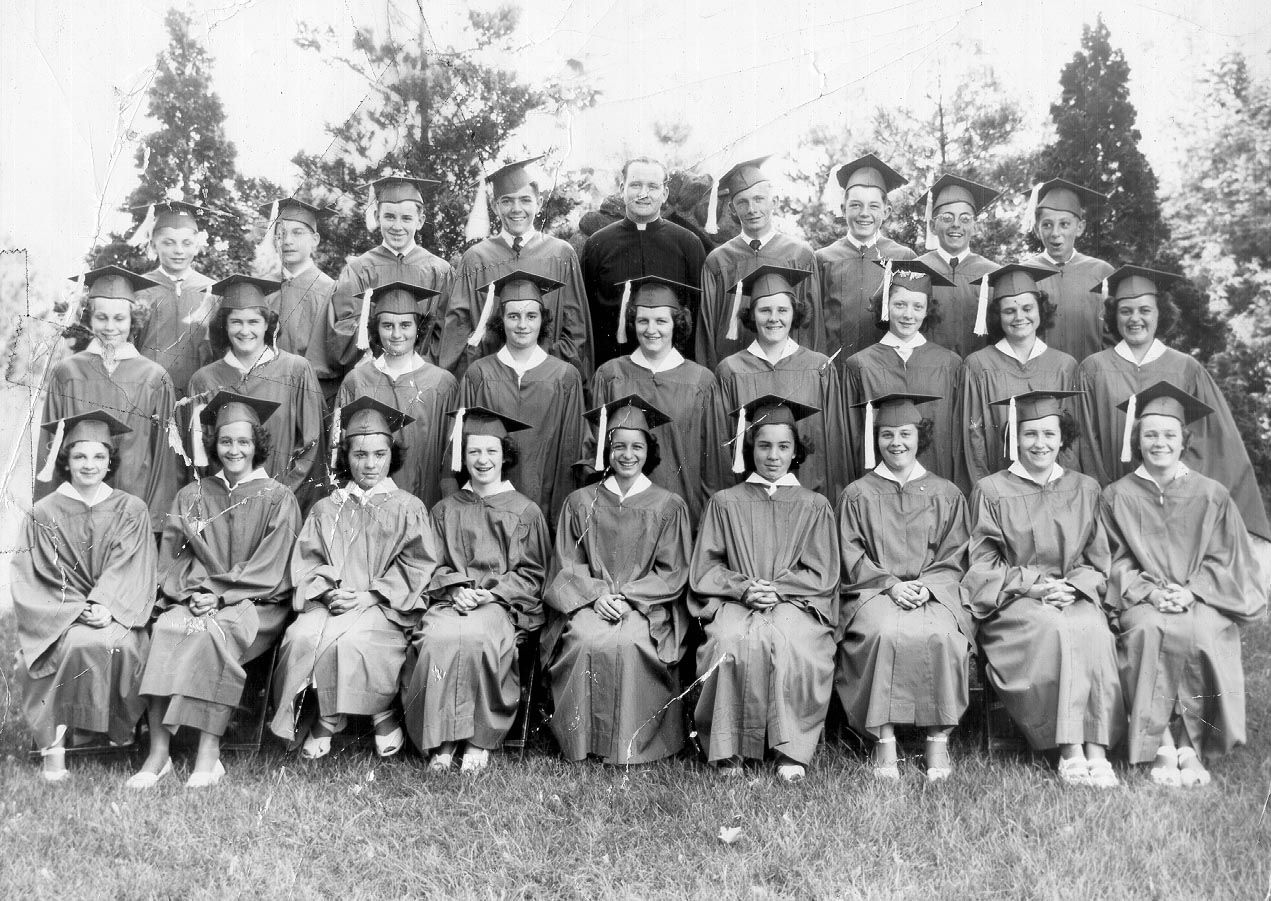 Black and white photo taken outdoors from 1949 of a group of young men and women in dark graduation gowns and caps with a man in a black priest's cassock and white collar