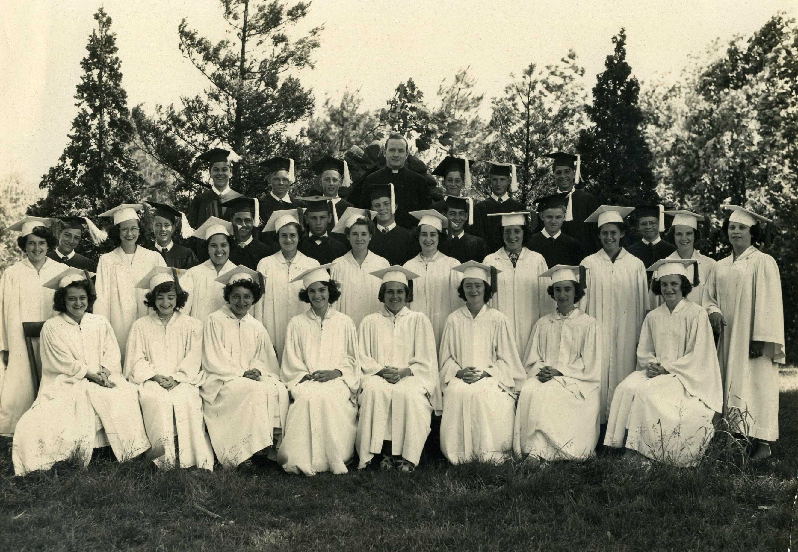 Black and white photo taken outdoors from 1950 of a group of young men in dark graduation robes and caps and young women in white graduation robes and caps, with a man in a black priest's cassock and white collar
