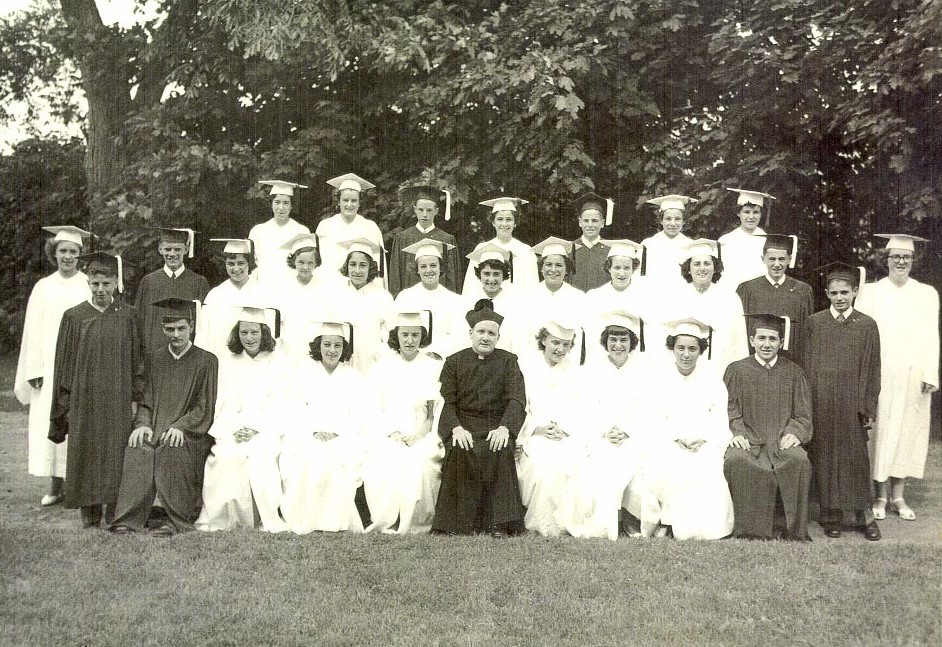 Black and white photo taken outdoors from 1951 of young men in dark graduation caps and gowns and young women in white graduation caps and gowns with a man in a black priest's cassock and white collar with black hat