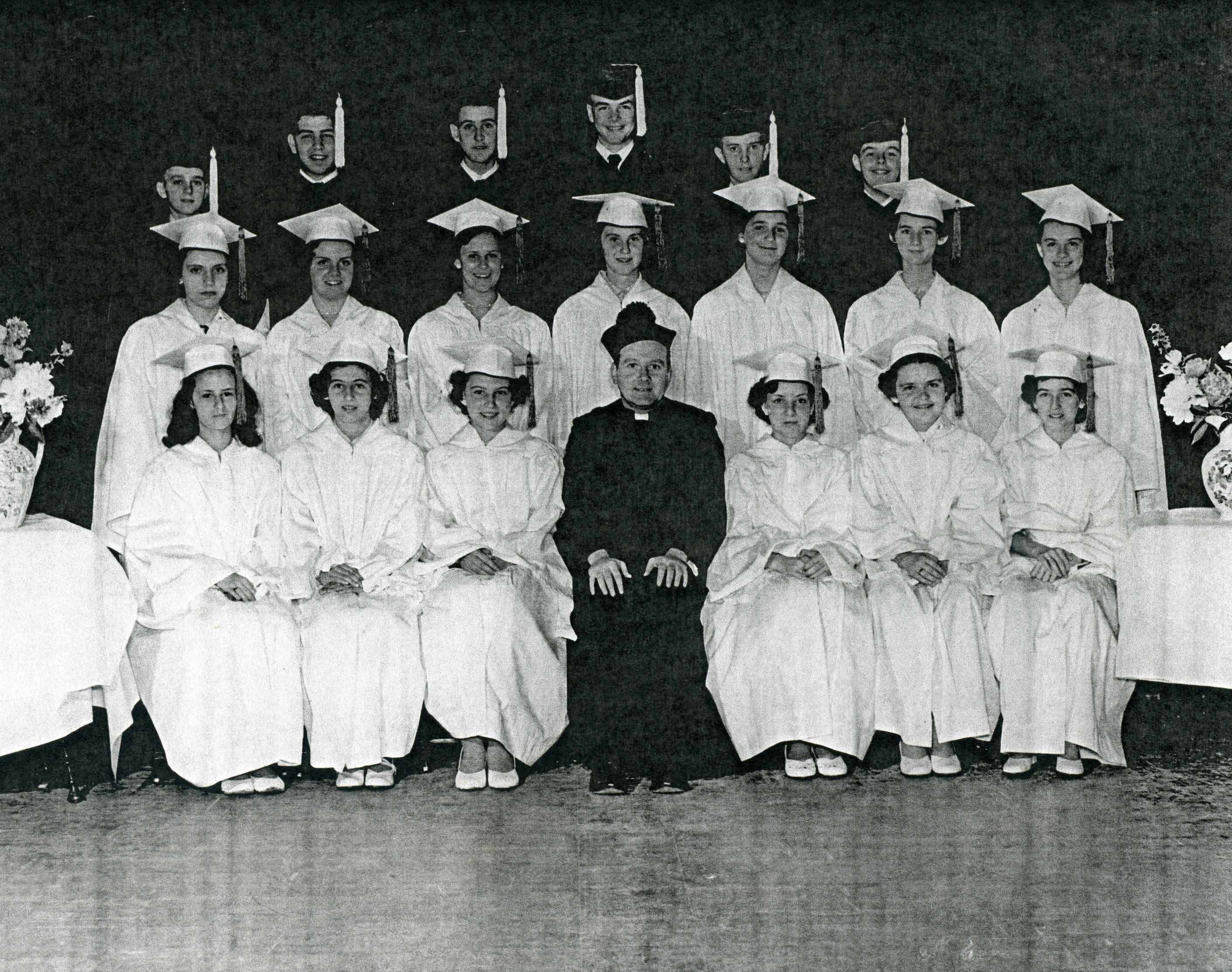 Black and white photo from 1953 of a group of young men in dark graduation caps and gowns and young women in white graduation caps and gowns with a man in a black priest's cassock with white collar and black hat