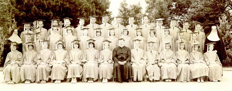 Black and white photo taken outdoors from 1960 of a group young men in dark graduation caps and gowns and young women in white graduation caps and gowns with a man in a black priest's cassock and white collar and two women in black nun's habits with white collars