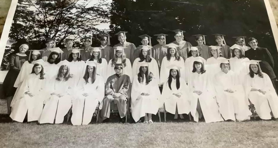 Black and white photo taken outdoors from 1971 of a group young men in graduation caps and gowns and young women in white graduation caps and gowns with a man in a black priest's cassock and white collar and a woman in a dark nun's habit and white collar