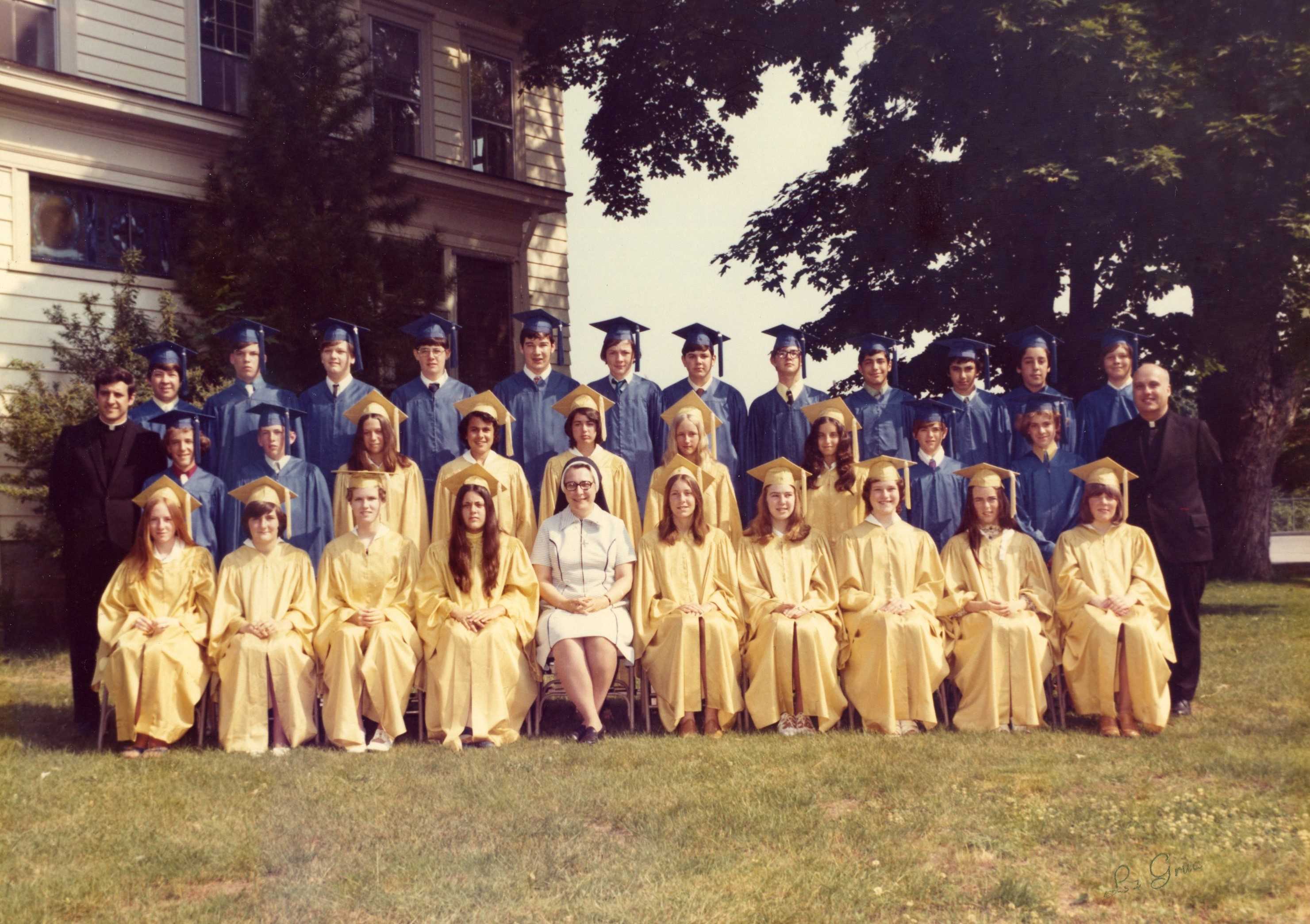 Photo from 1974 in front of building and large tree of a group young men in blue graduation caps and gowns and young women in yellow graduation caps and gowns with two men in a black priest suits and white collar and a woman in a white nun's habit and dark veil
