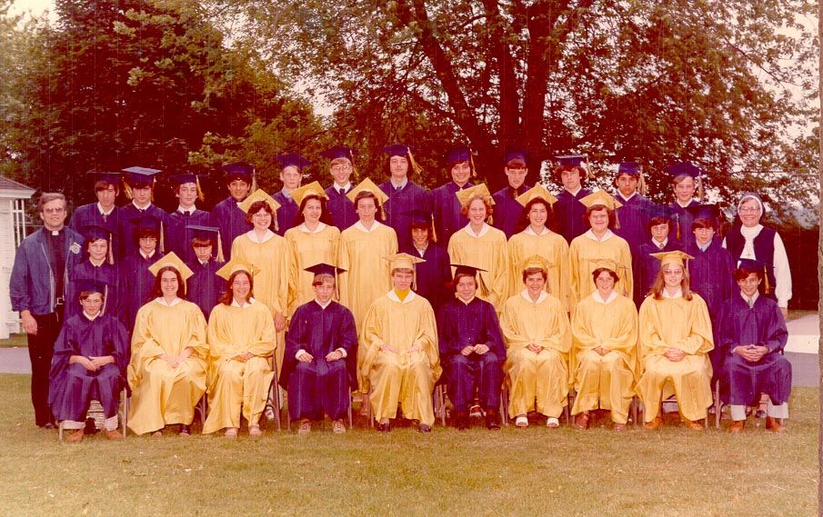 Photo from 1976 taken in front of a large tree of a group young men in blue graduation caps and gowns and young women in yellow graduation caps and gowns with a man in a black priest suit and white collar and a woman in a white and black nun's habit and dark veil