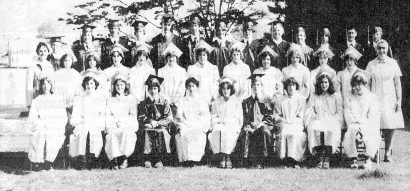 Black and white photo taken in front of a tree from 1978 of a group young men in graduation caps and gowns and young women in white graduation caps and gowns with a woman in a white nun's habit and dark veil and a woman in a dress