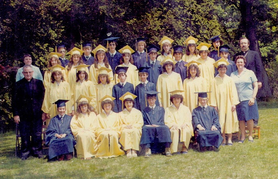 Photo from 1982 taken outdoors of a group young men in blue graduation caps and gowns and young women in yellow graduation caps and gowns with two men in black priest suits and white collars and two women in light blue shirts and dark skirts