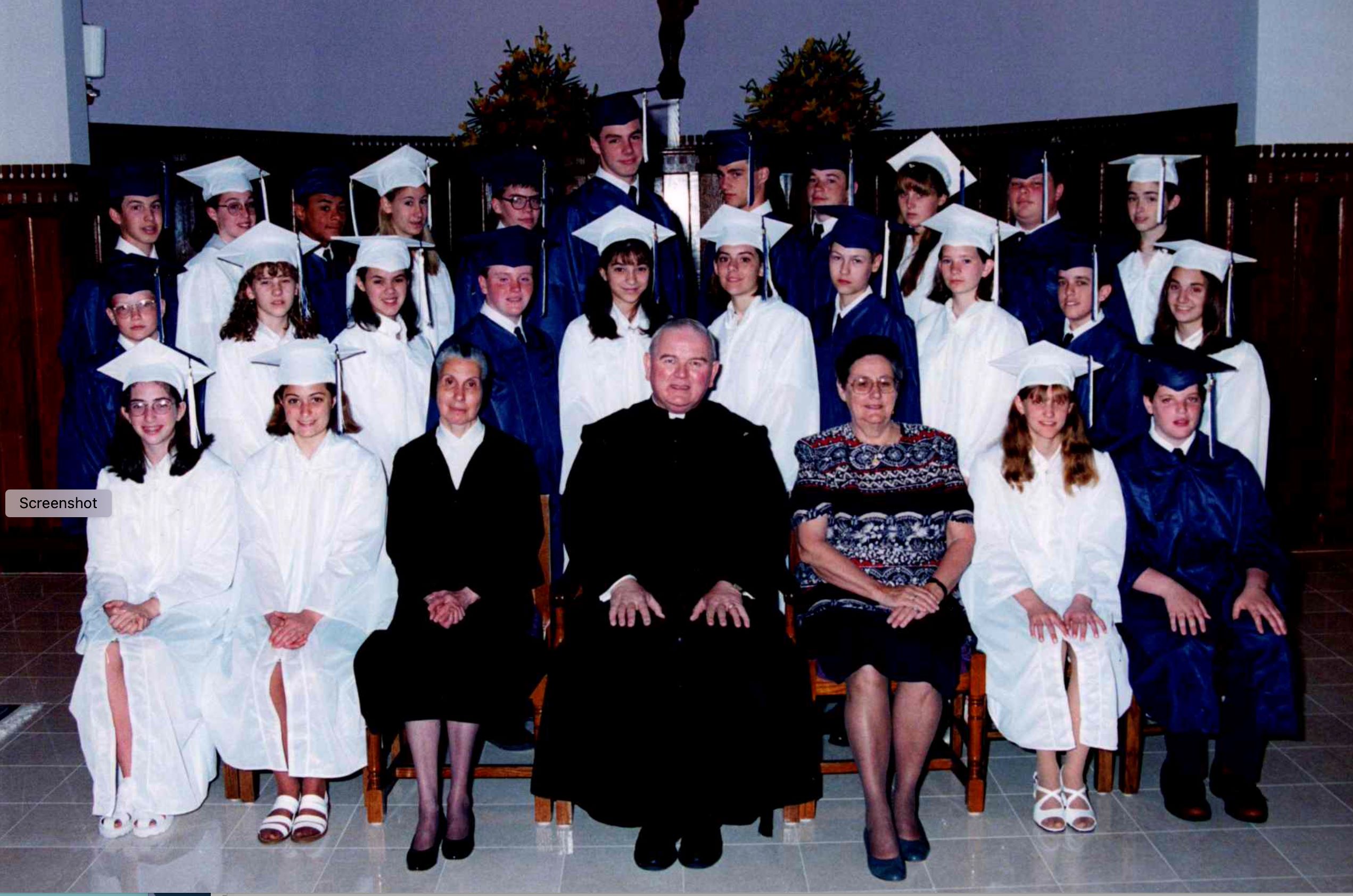 Photo from 1997 of a group young men in blue graduation caps and gowns and young women in white graduation caps and gowns with a man in a black priest suit and white collar and a woman in a dark nun's habit and dark veil