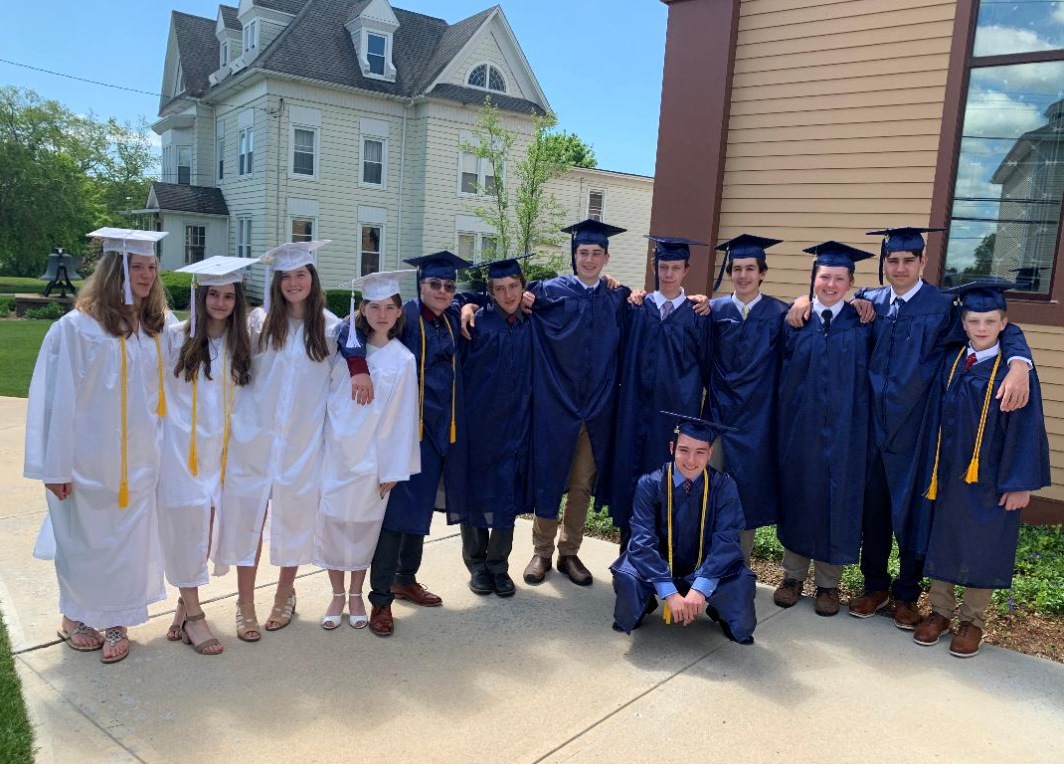 Photo from 2022 in front of a church of a group young men in blue graduation caps and gowns and young women in white graduation caps and gowns